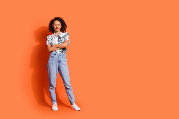 照片年轻迷人的女士棕色波浪头发穿着牛仔裤和夏威夷衬衫折叠手臂自信孤立的橙色背景图片下载