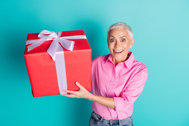 照片中漂亮的老妇人拿着大礼盒，穿着蓝绿色背景上孤立的粉色衬衫图片下载