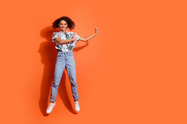 美国妇女全身长度的照片，在牛仔裤夏季衬衫跳跃空气和舞蹈与良好的共鸣孤立的橙色背景图片下载