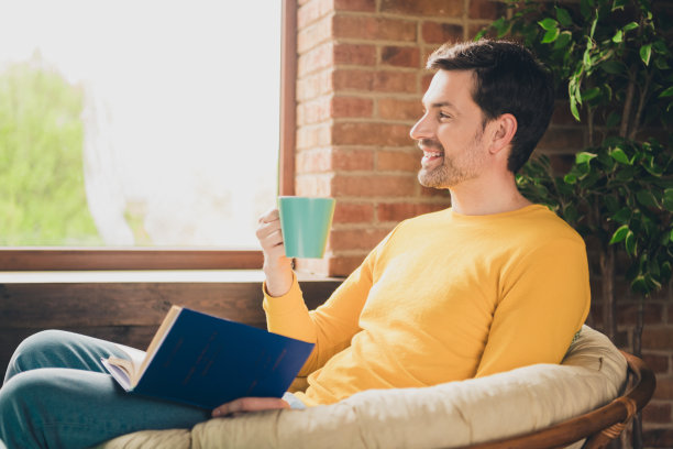英俊成熟男人的照片肖像坐在扶手椅喝茶看书穿着黄色的衣服舒适的日光家室内客厅图片下载