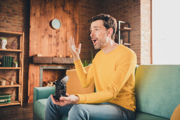 英俊成熟男人的照片肖像惊奇地玩电子游戏坐在沙发上穿着黄色的衣服舒适的日光家室内客厅图片下载