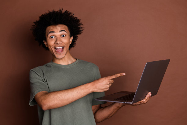 照片的年轻人在灰色t恤远程工作远程使用笔记本电脑指着显示器孤立的棕色背景图片下载