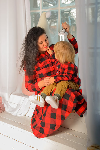 妈妈和儿子穿着相同的节日圣诞服装，坐在窗台上，拿着圣诞灯。家人看。圣诞奇迹。平安夜，母亲带着小儿子在家里望着窗外的照片。妈妈和孩子图片下载