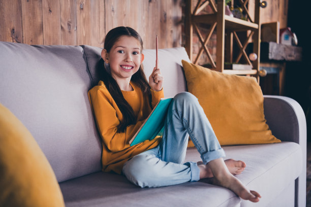 可爱小女孩的全尺寸照片坐在沙发上写笔记本尤里卡的想法穿着橙色的衣服舒适的日光家室内客厅图片下载