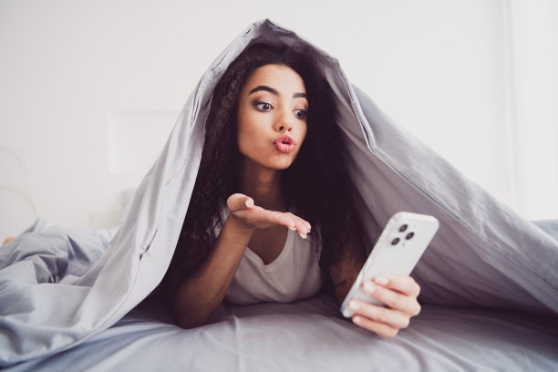 年轻女子躺在床上的照片盖着毯子送飞吻自拍照片穿着时尚的睡衣柔软舒适的卧室白色房间内部图片下载