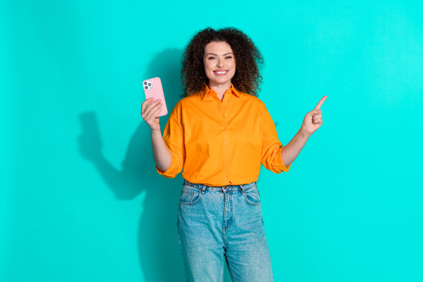 照片中，一个积极向上的女人，一头卷发，穿着橙色衬衫，手持智能手机，在蓝绿色的背景上指引空旷的空间图片下载