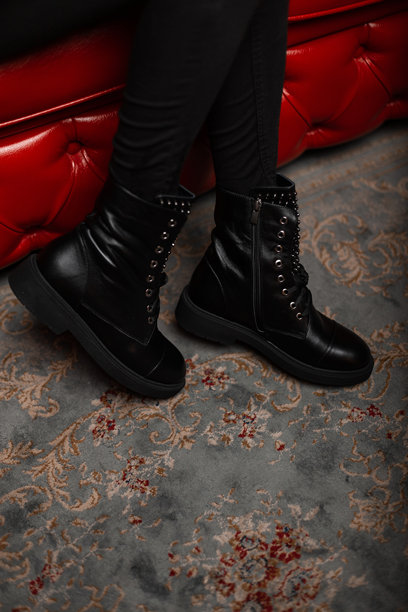 女性的腿在室内穿着时髦的黑色皮靴和绑带牛仔裤。时尚的年轻女子穿着时髦的鞋子坐在商店里别致的红色沙发上。秋春休闲风格。图片下载