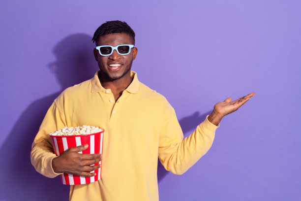 帅气的年轻人看电影爆米花hold点空穿着时尚的黄色服装孤立在紫色的背景图片下载