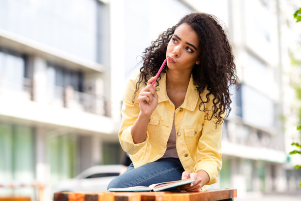 年轻的卷发女孩的照片，穿着黄色时髦夹克的学生在户外花时间拿着铅笔在抄写本上写笔记图片下载