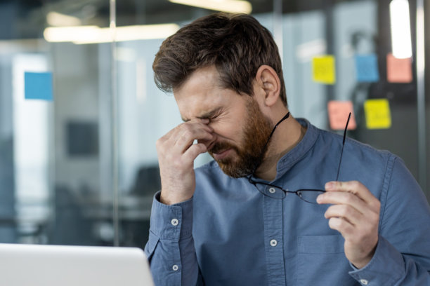 一个疲惫的年轻商人在办公室用笔记本电脑工作的特写照片，他拿着眼镜，因疲劳和超负荷而揉着眼睛图片下载