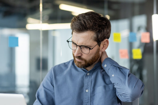 一个穿着蓝色衬衫的年轻人疲惫地坐在办公室里，用手按摩脖子的特写照片图片下载
