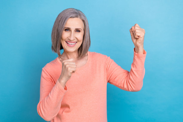有趣的退休妇女的照片，灰白的头发，穿着桃红色的时髦衬衫，举起拳头庆祝胜利，在蓝色的背景下购物图片下载