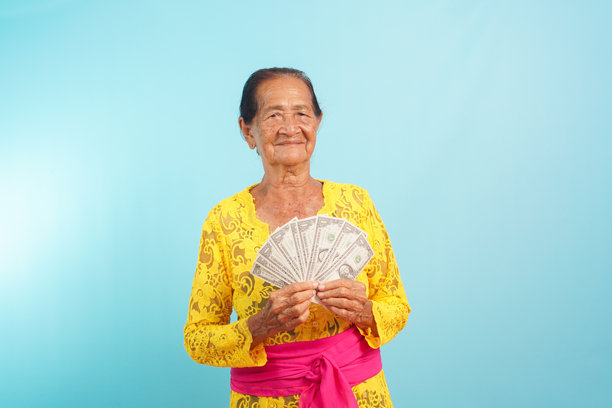一位身穿传统巴厘黄布的老妇人，手持蓝色背景的钞票扇子图片下载