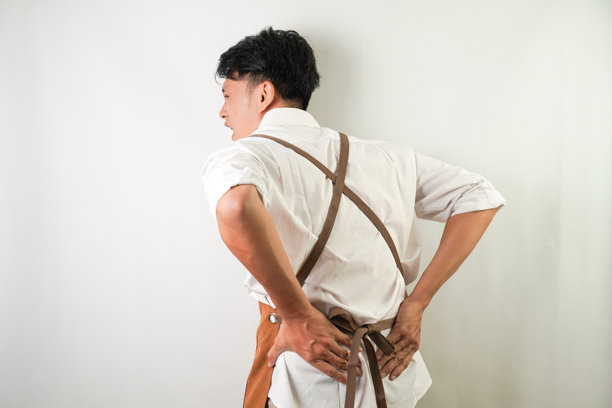 穿着卷起的白衬衫，系着棕色围裙的年轻亚洲人，正苦笑着腰肌肉疼痛。隔离在白色背景上。图片下载