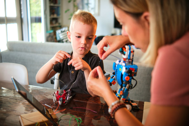 照片中，一位母亲正在帮助她年幼的儿子做一个色彩缤纷的机器人项目，强调在光线充足的室内环境中指导和教育的重要性。图片下载