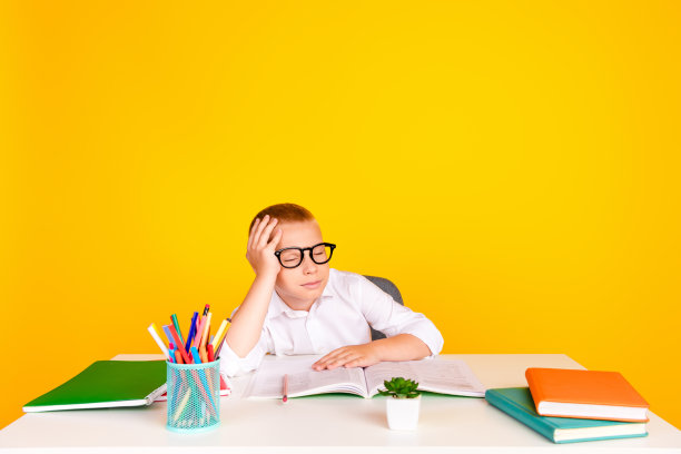 照片聪明迷人可爱困倦的男孩坐在书桌作业练习考试准备孤立的黄色背景图片下载