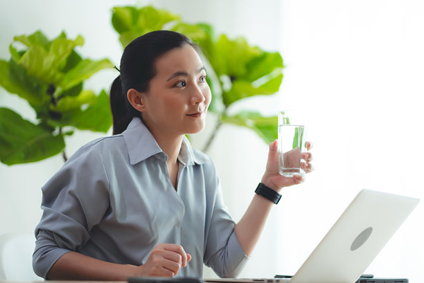 亚洲女性坐在家里办公室边喝水边用笔记本电脑休息。图片下载