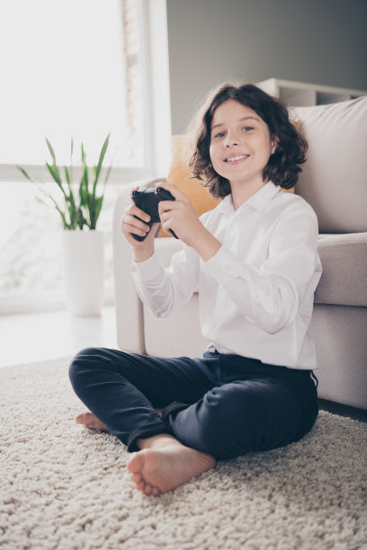 非常可爱的正面小男孩的照片，小学生坐在地板地毯上玩游戏，家里公寓房间室内室内图片下载