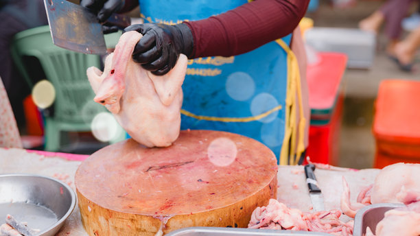 一个屠夫正在砧板上切鸡。故事发生在一个市场，背景中有几个人。屠夫戴着手套，围着围裙图片下载
