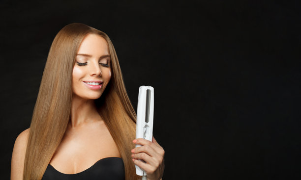 在黑色背景的工作室里，一个开朗的女人拿着直发器。护发、保健及年轻女模特用直发棒进行美发、造型及护发图片下载