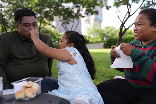 一个又大又胖的亚洲家庭，爸爸、妈妈、女儿在公园里野餐，他们坐着吃零食。他们坐着做作业，互相送花。他们很开心。图片下载