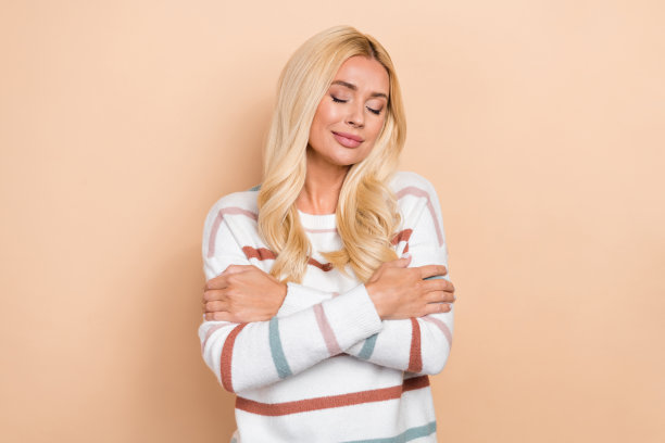 照片满意的高兴的女士积极闭上眼睛拥抱自己舒适的洗衣条纹毛衣新鲜的衣服隔离在米色背景图片下载