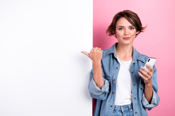 迷人可爱的女孩拿着智能手机的照片，手指空白的广告牌孤立在粉红色的背景图片下载