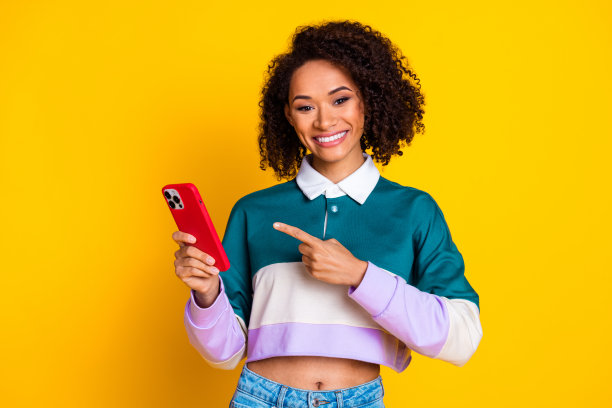 照片中，一个开朗的女人，一头卷发，穿着条纹上衣，手臂上拿着智能手机，背景是黄色图片下载