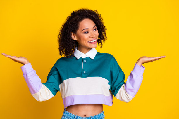 开朗可爱的卷发女人的照片，穿着条纹上衣，看着手臂上的物体，空白的空间孤立在黄色背景上图片下载