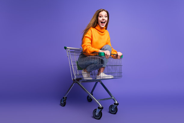 照片肖像金发十几岁的女孩在橙色高领毛衣和牛仔裤骑超市推车孤立的紫色背景图片下载