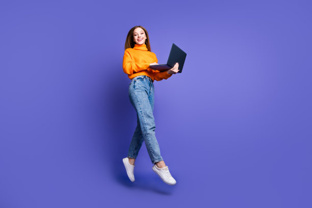 照片肖像金发开朗的年轻女孩在橙色高领毛衣和牛仔裤跳跃与电脑孤立的紫色背景图片下载