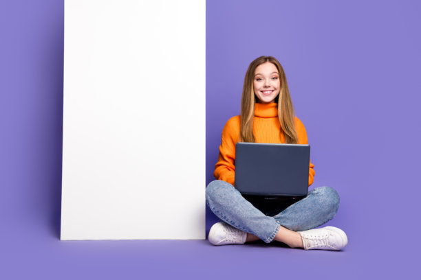 照片中，十几岁的女孩穿着橙色毛衣和牛仔裤，坐在地板上，笔记本电脑靠近手机模板板，孤立在紫色背景上图片下载