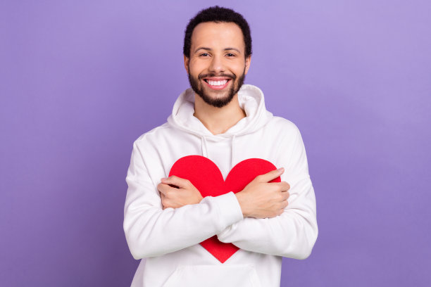 照片中开朗率真的男子笑容满面，双臂抱抱纸红色的心，孤立在紫罗兰色的背景上图片下载