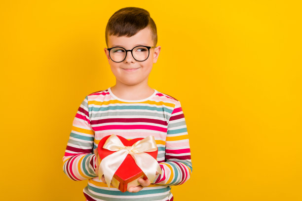 照片中开朗狡猾的男孩穿着时髦的衣服，拿着礼盒心形，看起来很感兴趣，空白的空间孤立在黄色的背景上图片下载