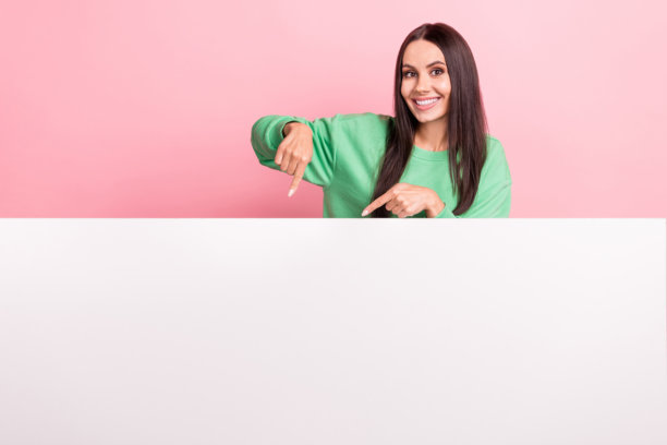 照片中，开朗、积极、可爱的女人穿着绿色的运动衫，看着粉红色背景上的广告牌空白图片下载