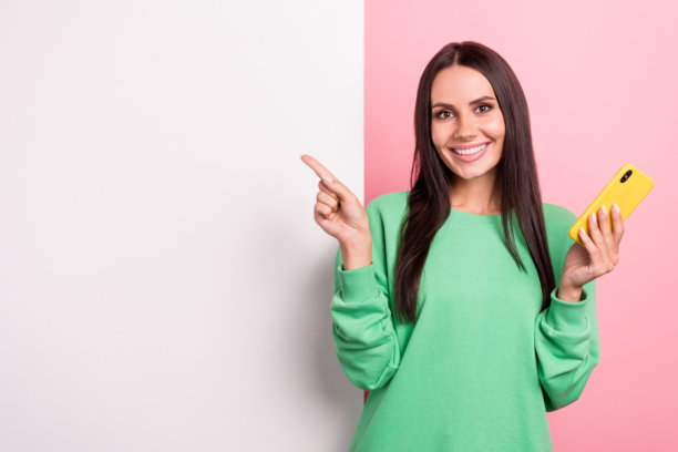 照片微笑的漂亮的年轻女士穿着绿色套头衫直接手指纸白板拿电话推荐购物孤立在粉红色的背景图片下载