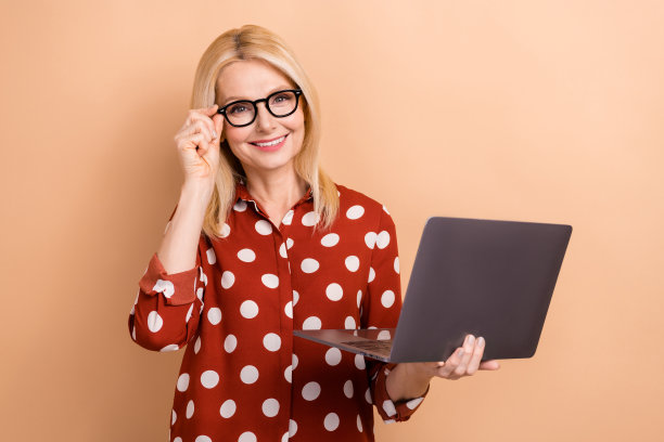 照片中，开朗的资深女性梳着波波头，穿着带点的衬衫，拿着笔记本电脑，手指触摸，眼镜孤立在柔和的色彩背景上图片下载