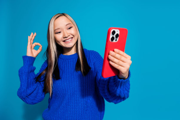 年轻女孩的照片肖像，穿着时尚的针织毛衣，用手机博主展示了蓝色背景上孤立的ok符号图片下载