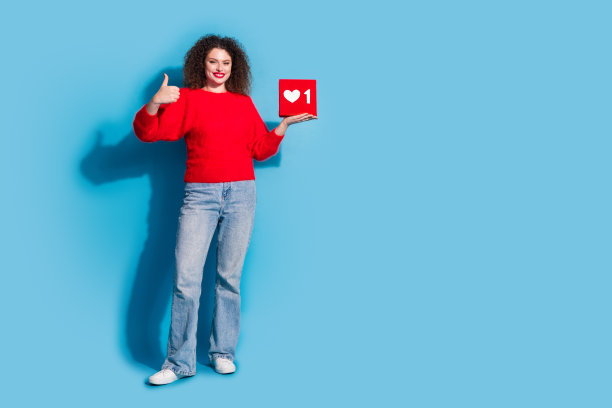 可爱的年轻女士的全身照片，拇指向上拿着社交媒体图标，穿着时尚的红色服装，与蓝色背景分开图片下载