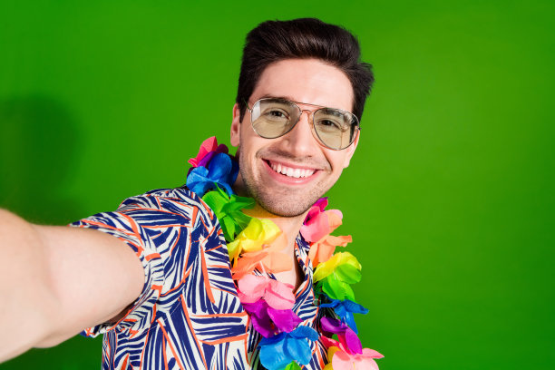 照片上，一个积极向上的人，穿着刚毛印花衬衫，戴着眼镜项链，在绿色背景下用智能手机自拍图片下载