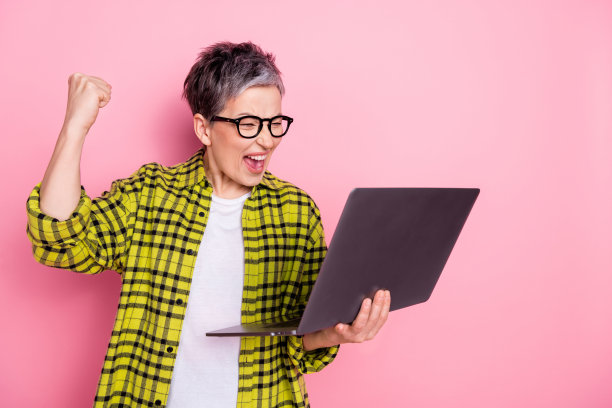 照片中漂亮的老年妇女举起拳头，使用笔记本电脑，格子衬衫孤立在粉红色背景上图片下载