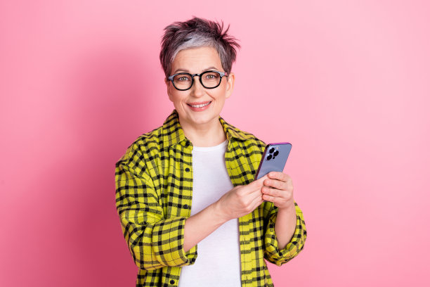漂亮的老年妇女的照片使用手机格子衬衫孤立的粉红色背景图片下载