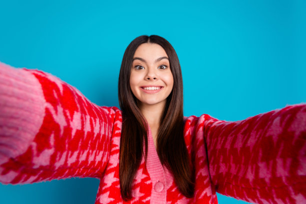 照片中，一个长着牙齿、笑容满面的漂亮女人，直发，穿着针织衫，开襟羊毛衫，在蓝色的背景下自拍图片下载