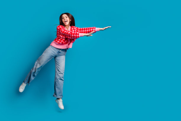 全尺寸照片的好心情可爱的女人穿着针织衫，开襟羊毛衫牛仔裤飞到空旷的空间孤立的蓝色背景图片下载