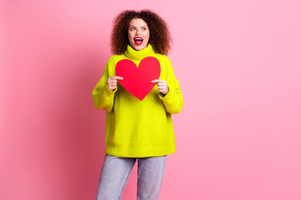照片中可爱的年轻女士抱着大红色的心，看起来很惊讶，空白的空间穿着时髦的黄色衣服，孤立在粉红色的背景上图片下载