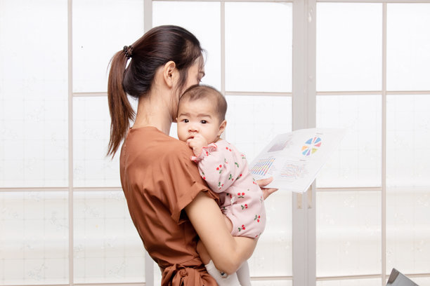 美丽的亚洲母亲抱着她蹒跚学步的女儿一边看书，新生儿穿着粉红色的棉软纺织品樱桃图案。穿着棕色连体衣的女人。图片下载