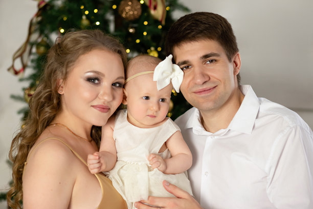 一个美丽家庭的特写照片，父亲和母亲抱着一个小女儿，背景是一棵为圣诞节和新年装饰的圣诞树。幸福家庭理念图片下载