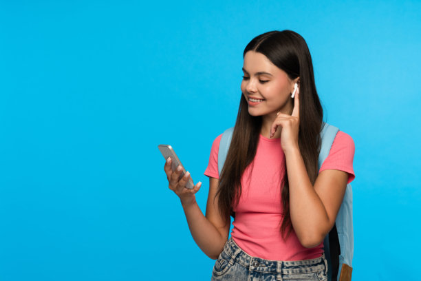 开朗的女孩拿着包拿着智能手机，戴着无线耳机，与世隔绝的蓝色背景图片下载