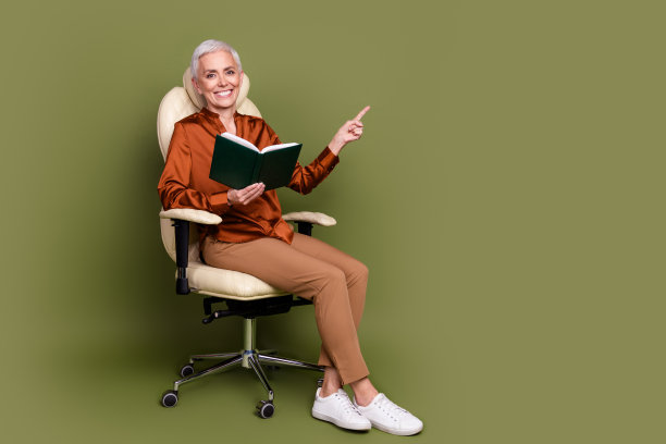 老年妇女的全身照片坐在扶手椅上看书点空空间穿着棕色正装孤立在卡其色背景图片下载