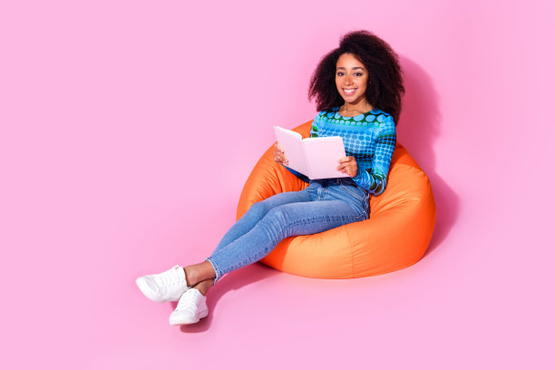 漂亮的年轻女孩的全尺寸照片，坐在橙色豆袋看书，穿时髦的蓝色衣服孤立在粉红色的背景图片下载
